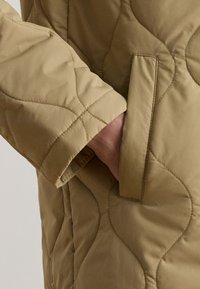 Hand in der Tasche einer hellbraunen gesteppte Jacke mit gebogenem Steppmuster und sichtbarer Taschennaht.