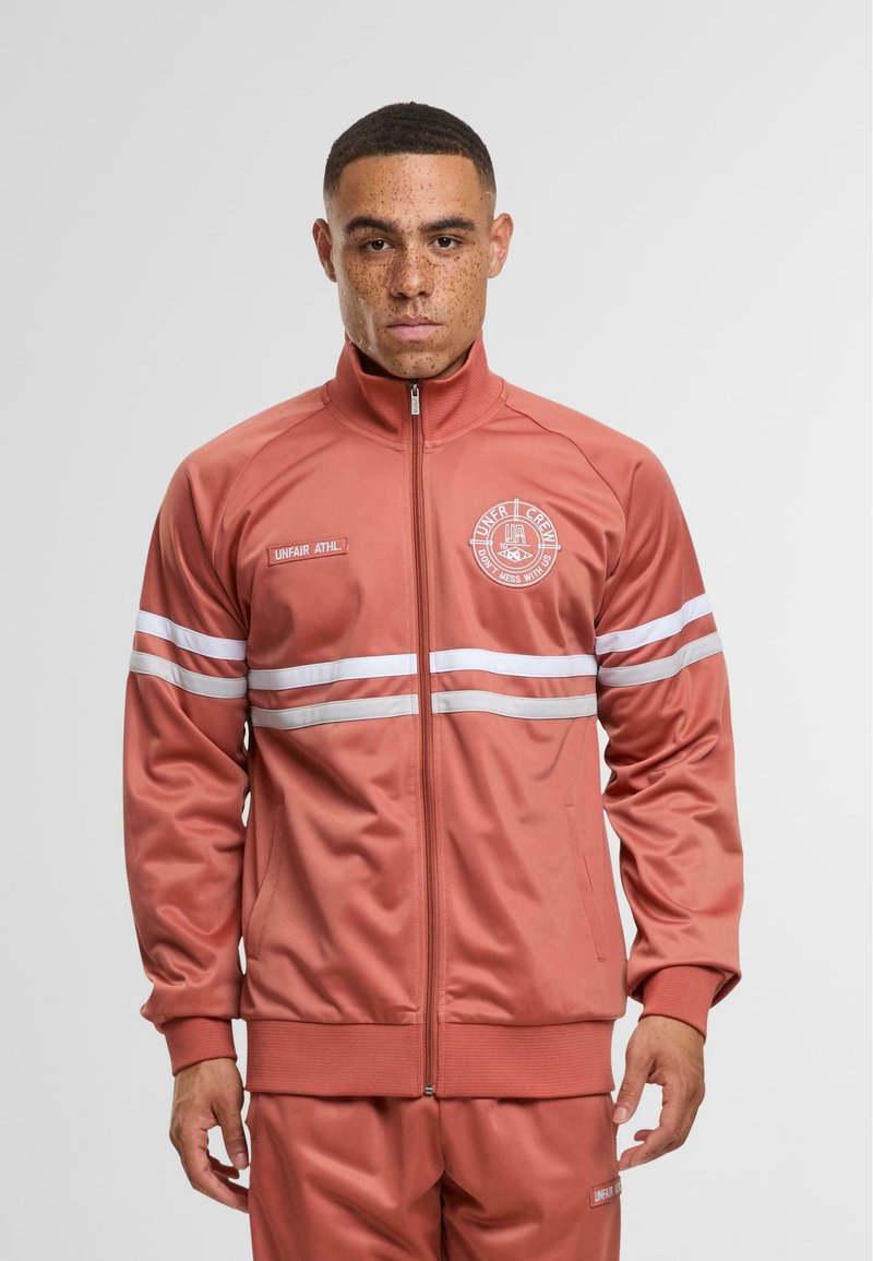 Zip-up trackjakke i støvet rose med hvide striber, ribbede manchetter og et logo-patch på brystet. Lavet af glat, letvægtsstof.