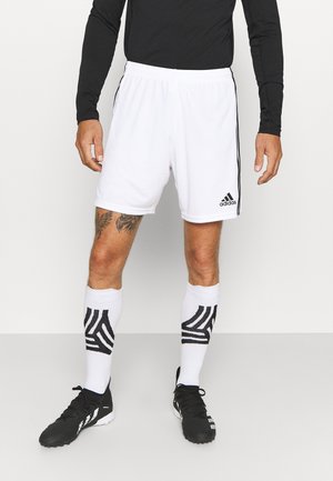 Mann trägt weiße Adidas-Fußballshorts, weiße Socken mit schwarzen Mustern und schwarze Fußballschuhe, steht vor einem einfarbigen Hintergrund.