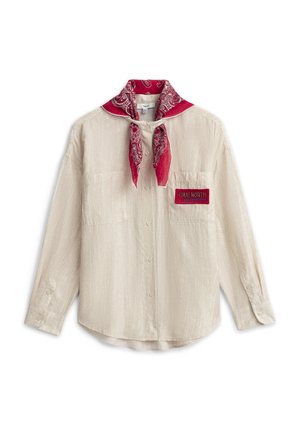 Chemise beige à boutons avec deux poches poitrine, un écusson rouge marqué "True North" et un bandana rouge à motifs noué autour du col.