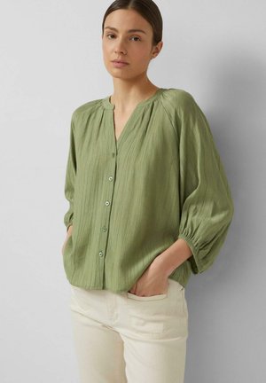 Woman wearing a loose, light green button-up blouse with three-quarter sleeves and cream-colored pants, standing with hands in pockets.
