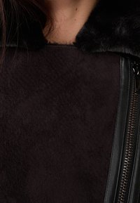 Sort ruskindsjakke med et falsk pelskrave, struktureret stof og en sidelukning med metalhardware.