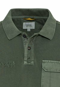 Olijfgroene polo met een gestructureerde kraag, twee knopen en een borstzak. Bevat geborduurde tekst "EXPLORERS 1977."