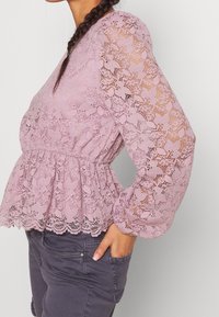 Blusa in pizzo rosa chiaro con maniche lunghe, design a peplum e motivi floreali intricati. Presenta una vita cintrata e un tessuto testurizzato.