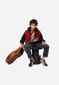 Rode puffer vest over een zwarte hoodie, graphic t-shirt, grijze jeans. Zwart en tan sneakers. Zittend op een zwart gespikkelde kruk, met een gitaar in de hand.