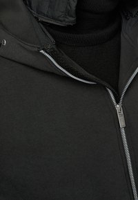 Black zip-up hoodie with a smooth texture, featuring a silver zipper and a lined collar. Interior layer in a ribbed black fabric.