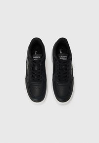 Ténis de pele preta com pontas redondas e atacadores planos. Apresenta detalhes perfurados na parte frontal e uma sola de borracha branca. Logótipo da Lacoste na lateral.