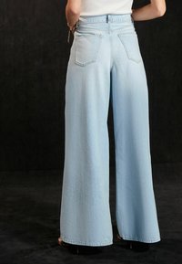Jean large bleu clair en denim, avec une taille haute, deux poches arrière et des détails cousus, reposant au-dessus de talons marron.