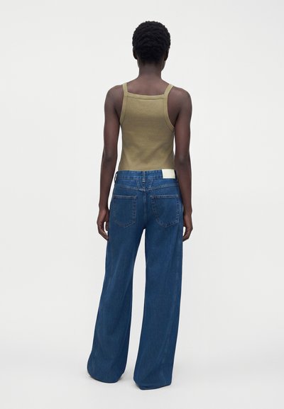 Personne debout, de dos, portant un débardeur vert olive et un jean bleu large sur un fond uni.
