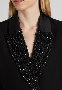 Blazer nero con revers in satin, impreziosito da perline nere e cristalli. Il colletto presenta dettagli angolari, creando un contrasto di texture.