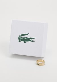 Zlatni prsten s logotipom zelenog krokodila, prikazan pored bijele kutije koja sadrži isti logotip u zelenoj boji. Kutija ima teksturiranu površinu.
