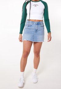 Světle modrá džínsová mini sukně s roztrhaným lemem, kombinovaná s bílou košilí s dlouhým rukávem a zelenými rukávy, na které je logo. Doplněno o bílé tenisky.
