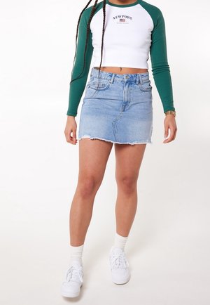 Mini gonna di jeans azzurro chiaro con orlo sfrangiato, abbinata a una camicia bianca a maniche lunghe con maniche verdi e un logo. Indossata con sneakers bianche.