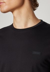 Sort bomulds T-shirt med en glat tekstur, rund hals og et lille sort logo-patch på venstre side.
