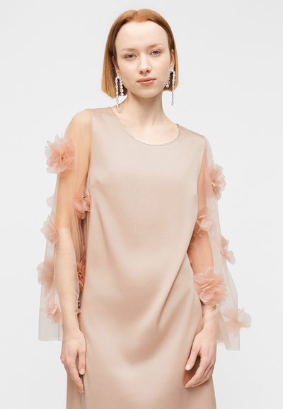 Νέα γυναίκα με κοντά κόκκινα μαλλιά, που φοράει μπεζ αμάνικο φόρεμα με διαφανείς μανίκια διακοσμημένα με υφασμάτινα λουλούδια και μακριά κρυστάλλινα σκουλαρίκια.