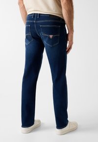 Uomo che indossa jeans blu scuro con tasche posteriori cucite e sneaker bianche, in piedi contro uno sfondo chiaro e uniforme.
