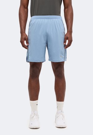 Homme portant un short de sport bleu clair, des chaussettes blanches Puma et des baskets blanches, debout, les bras détendus le long du corps, devant un fond neutre.