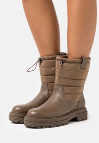 Bottines imperméables brun avec une partie inférieure en cuir texturé et une partie supérieure en nylon matelassé, dotées d'une fermeture à cordon et d'une semelle épaisse.