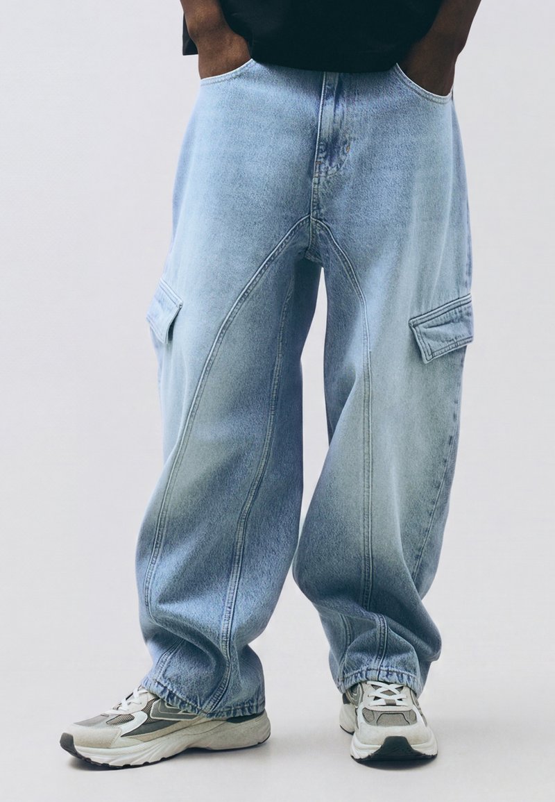 Personne portant un jean cargo bleu clair ample et des baskets blanc-gris, les mains dans les poches sur un fond uni.