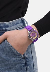 Paarse siliconen horloge met ronde wijzerplaat, zilveren accenten en grote gele cijfers. Draagt om een hand die in een denim zak rust.