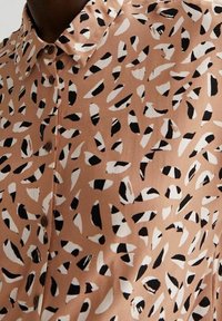 Camicia marrone con bottoni realizzata in tessuto liscio, caratterizzata da un motivo sparso di forme nere e bianche, con colletto classico e sfondo beige.