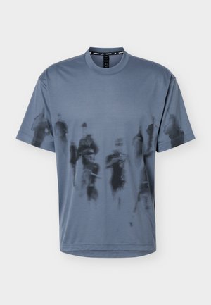 Hombre con cabello corto que lleva una camiseta gris de gran tamaño con patrones abstractos negros y pantalones azules, de pie frente a un fondo liso.