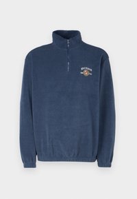 Marinblå fleece-pullover med kvartdragkedja, ribbade ärmslut och en logotyp på bröstet, som visar en mjuk textur och avslappnad passform.