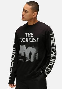 Svart långärmad t-shirt med en grå grafisk design och texten "THE EXORCIST" på framsidan och ärmen. Tillverkad av mjukt tyg.