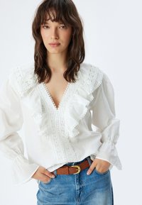 Witte blouse met ruches van kant, v-hals en lange mouwen. Gecombineerd met een bruine riem en spijkerbroek. Soepele textuur.