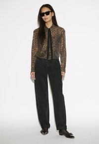 Doorzichtig blouse met luipaardprint en een zwarte strik, gecombineerd met hoge zwarte broek met lichte versieringen en zwarte instapschoenen.