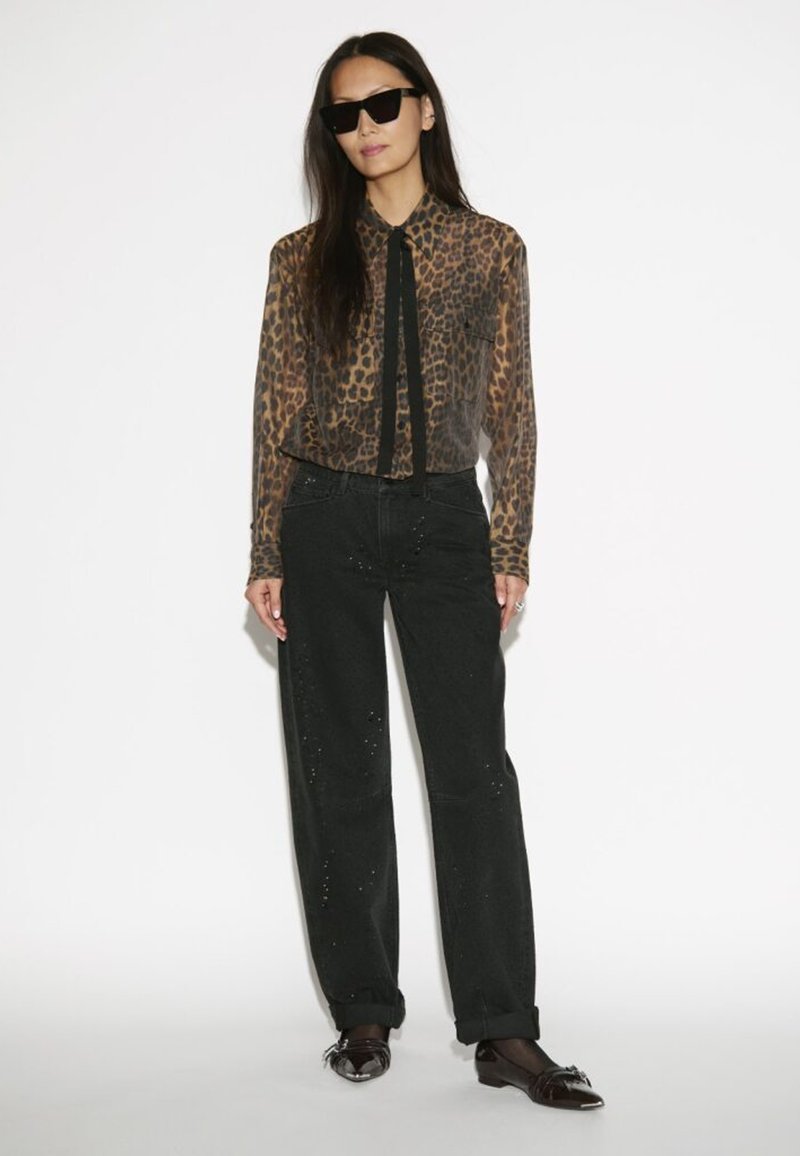 Doorzichtig blouse met luipaardprint en een zwarte strik, gecombineerd met hoge zwarte broek met lichte versieringen en zwarte instapschoenen.