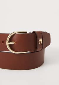 Brown leather belt with a gold-tone circular buckle and embossed logo detail. Smooth texture with evenly spaced belt holes.