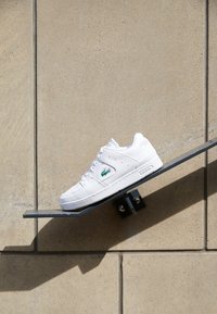 Bílé tenisky s hladkým koženým svrškem, perforovanou špičkou a zeleným logem. Mají plochou podrážku a zavazování na tkaničky. Zobrazené na nakloněné polici.