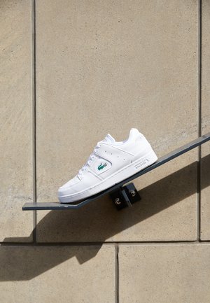 Baskets blanches avec une tige en cuir lisse, une boîte à orteils perforée et un logo vert. Présentent une semelle plate et un système de laçage. Exposées sur une étagère inclinée.