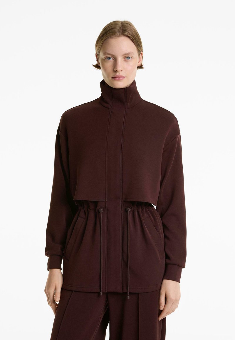 Vrouw met kort haar in een donkergbruine hoge kraag ritsjack met een trekkoord in de taille en bijpassende broek tegen een witte achtergrond.