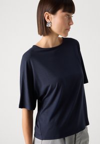 T-shirt blu navy a maniche corte, con scollatura rotonda, texture liscia, vestibilità rilassata, abbinato a orecchini d'argento, indossato con pantaloni chiari.