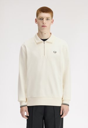 Jeune homme aux cheveux roux courts portant un sweat-shirt crème à demi-fermeture éclair avec un petit logo de couronne de laurier noire sur la poitrine.