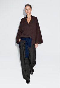 Brązowa koszula z guzikami w luźnym kroju, połączona z ciemnymi spodniami w prążki oraz granatowym swetrem związanym w talii. Gładka tekstura, swobodny wygląd.