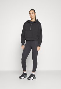 Czarna krótka bluza z kapturem, z zamkiem błyskawicznym na ćwierć długości i przednią kieszenią, w połączeniu z czarnymi legginsami i czarnymi butami sportowymi z białymi akcentami.