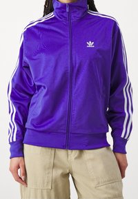 Lila polyester tracksuitjacka med hög krage, frontdragkedja och vita tre-stripes-accenter på ärmarna. Adidas-logotypen syns.