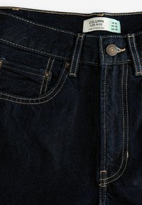 Vysoké temně modré džíny z denimu s viditelnými švy, předním knoflíkem a dvěma předními kapsami. Značka je přišita na opasku.