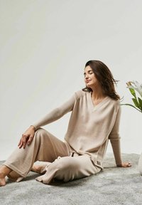 Beige loungewear set made of soft fabric, featuring a V-neck top with long ribbed sleeves and relaxed-fit pants, sitting on a textured surface.