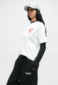 Γυναίκα με άσπρο καπέλο, άσπρη μπλούζα T-shirt με κόκκινα χείλη, μαύρα μανίκια κάτω από αυτήν και μαύρες φόρμες Jordan, με τα χέρια στις τσέπες.