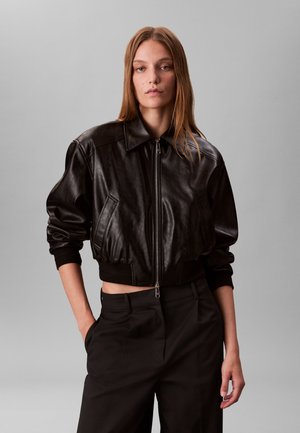 Junge Frau mit langen braunen Haaren, die eine schwarze kurze Lederjacke und schwarze Hose trägt, steht mit einer Hand in der Tasche vor grauem Hintergrund.
