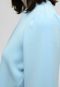 Blusa azul claro con un diseño estructurado. Presenta mangas abullonadas y una textura suave, destacando el corte holgado y el aspecto elegante.