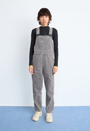 Grey denim overalls with adjustable straps, featuring a front pocket. Worn over a black long-sleeve top, complemented by beige shoes.