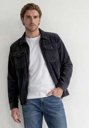 Mand med lyst brunt hår iført en sort manchesterjakke, hvid T-shirt og blå jeans, står op ad en hvid væg.