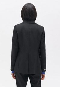 Blazer negro con un diseño ajustado, textura suave y solapas clásicas. Presenta acentos de botones plateados en las mangas y una silueta estructurada.