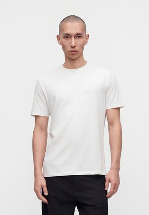 Homme à la tête rasée portant un t-shirt blanc uni à manches courtes et un pantalon noir, debout devant un fond blanc uni.