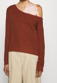 Brick red off-shoulder sweater with ribbed texture and long sleeves, paired with light-colored, loose-fitting pants.